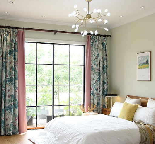 Modern bedroom with floor-to-ceiling custom printed curtains in pink and green leaf patterns, styled with a white bed and gold chandelier, perfect for printed curtains for the bedroom.